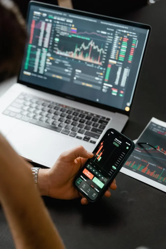 pexels-photo-7567228 Person analyzing stock market data on a smartphone with a laptop displaying financial graphs in the background.